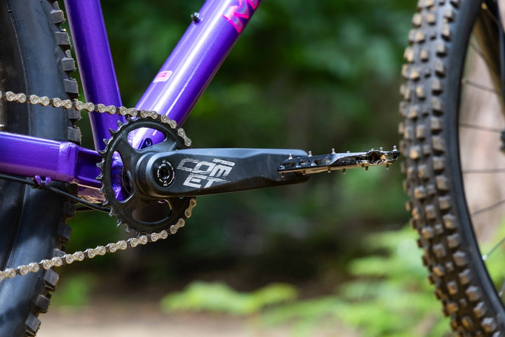 Close-up of a purple bicycle with a chain and gear system, blurred green background
