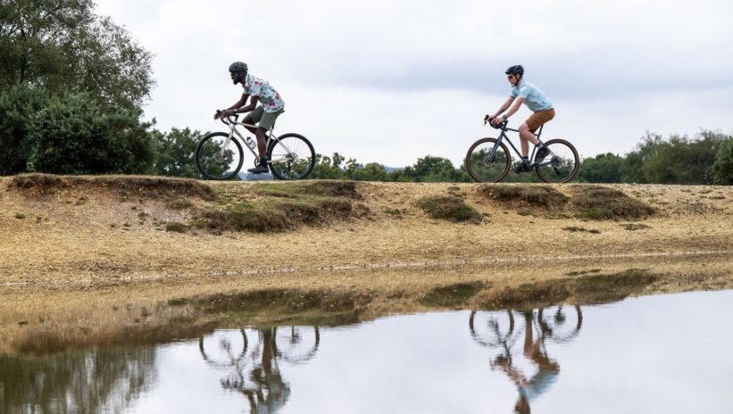 Two cyclists riding on a dirt path near a body of water with trees in the background.